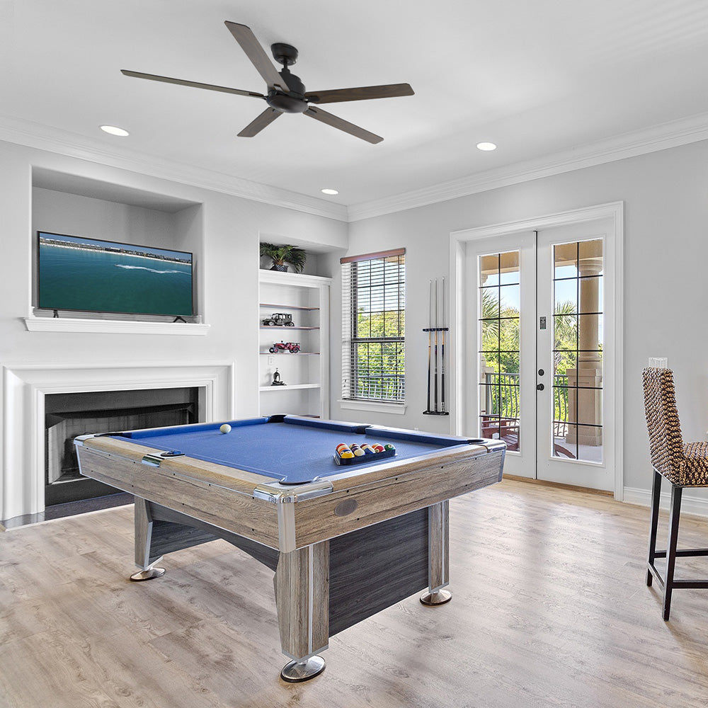 A spacious game room featuring a light wood-finish 60-inch Harvil Beachcomber pool table with a blue felt playing surface. A black Smafan or Carro modern, five-blade ceiling fan is mounted on the white ceiling. The room has light-colored wood flooring, a fireplace below a built-in TV, and glass French doors leading to a balcony. 
