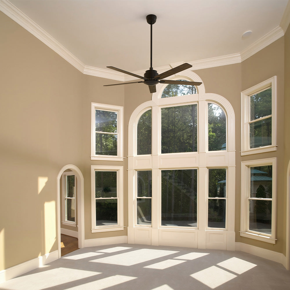 An interior view of a room with tall, arched bay windows, featuring a black ceiling fan mounted on a long downrod. The room has light tan walls and a light-colored carpet, with strong shadows cast across the floor by the abundant sunlight coming through the windows. The fan is consistent with a Smafan Warwick 60-inch model, which is typically a black ceiling fan without a light. 