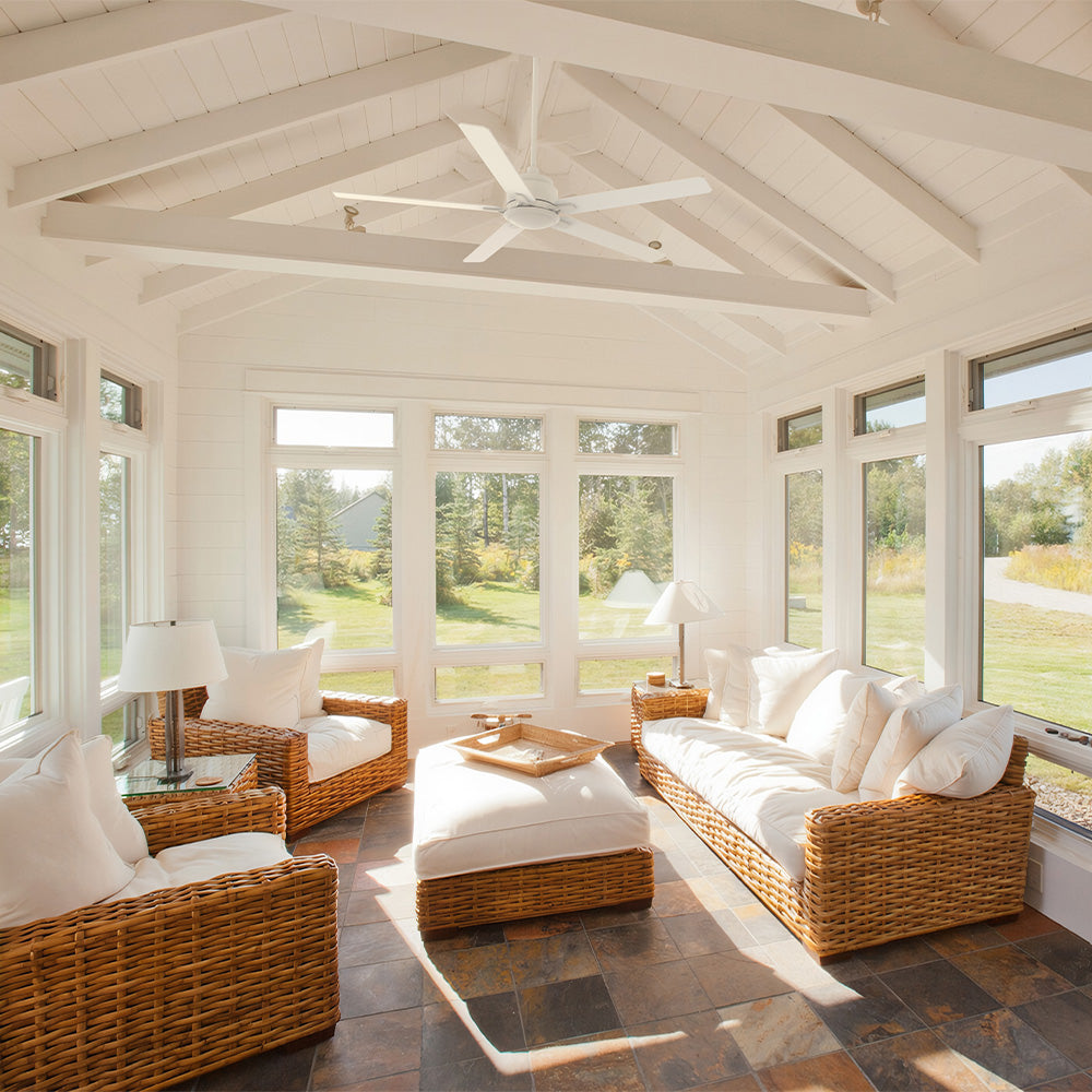 A spacious, sunlit sunroom features wicker furniture with white cushions and two glass-topped end tables on a dark slate or stone tile floor. Smafan Warwick 60-inch white ceiling fan without light, is mounted on a white painted, exposed-beam vaulted ceiling.  