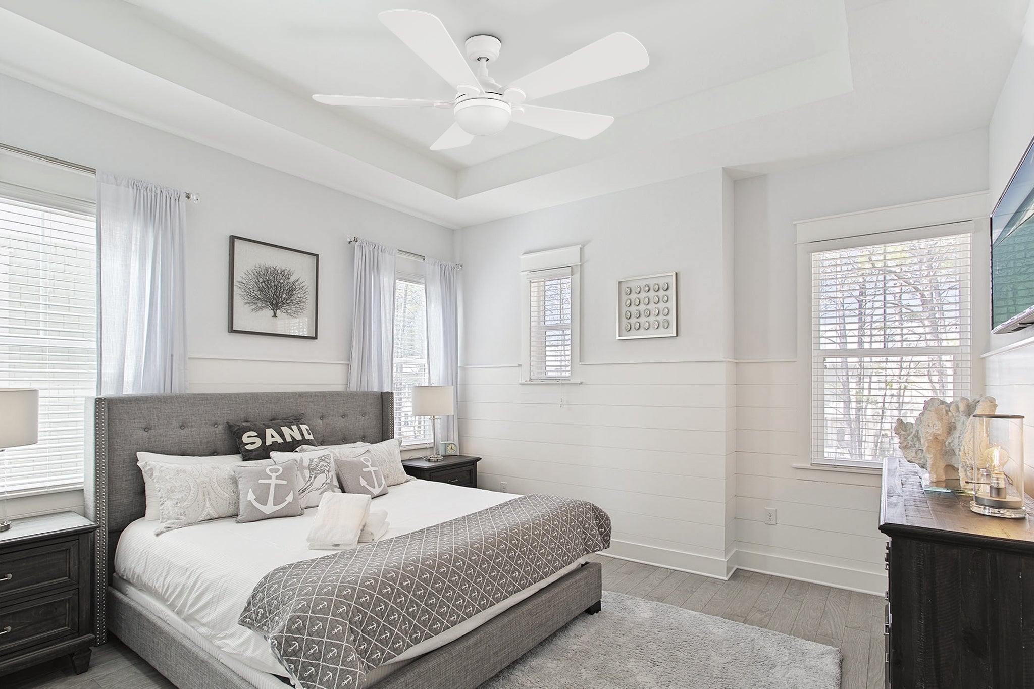 A bright modern bedroom with white walls, a tall tufted gray upholstered bed, and white bedding accented by gray pillows labeled “SAND” and an anchor design. Matching dark wood nightstands with lamps sit on each side, and a soft gray shag rug covers the floor. A white Smafan Lille 52-inch ceiling fan with LED light hangs from a tray ceiling, complementing the shiplap-style walls and natural light streaming through the windows with sheer curtains.
