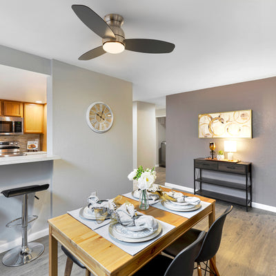 A modern dining area featuring a wooden dining table set for four, with two dark chairs visible. Above the table, a Trendsetter 48-inch low profile smart ceiling fan with LED light and remote control in a brushed nickel finish is installed. The fan has three dark gray blades and a central integrated LED light. 