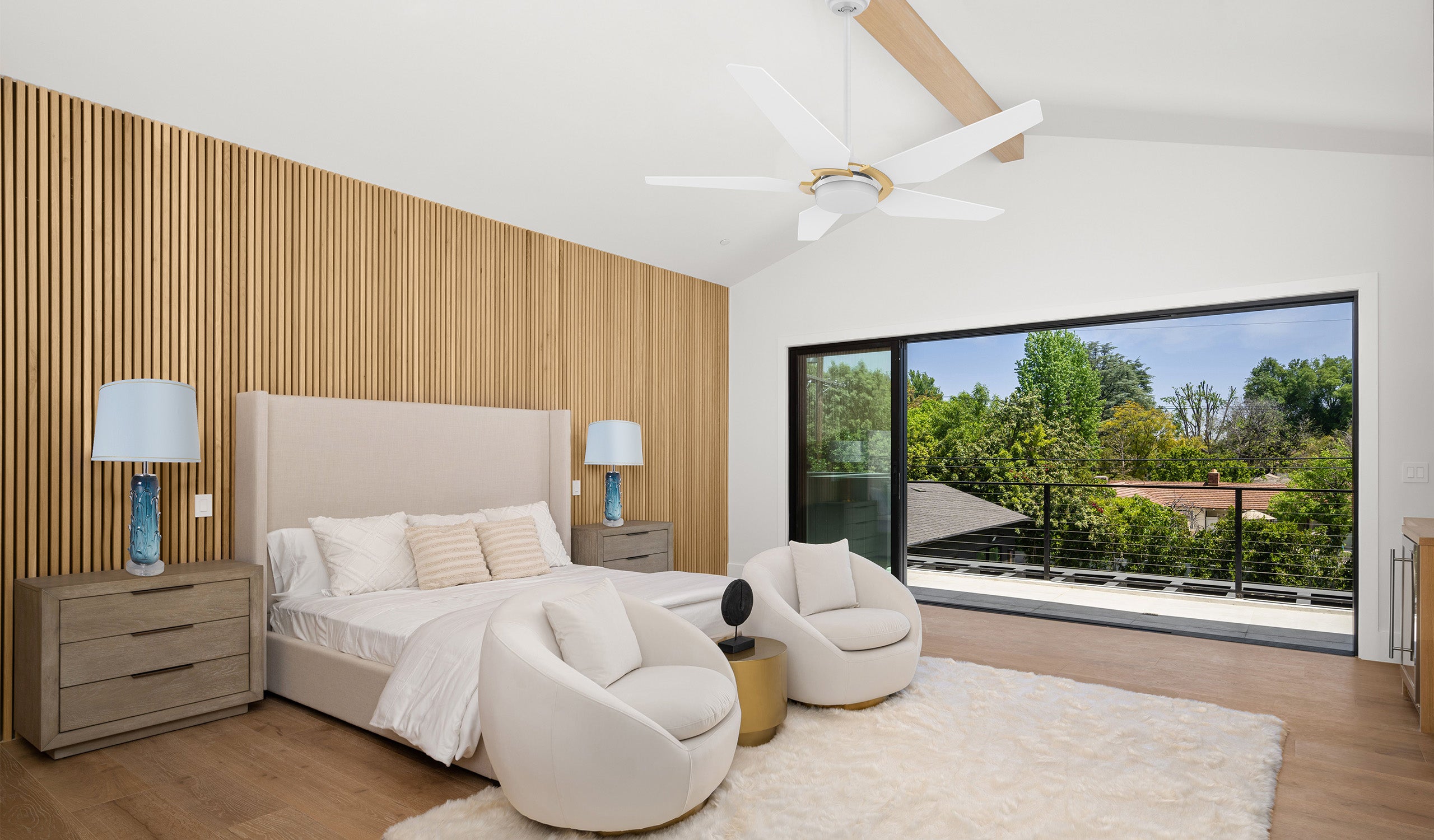 Modern minimalist bedroom featuring a sleek white ceiling fan with LED light and gold accents, a textured wooden accent wall, and a large sliding glass door leading to a balcony with lush greenery.  The room is furnished with a neutral upholstered bed, two contemporary white lounge chairs, a plush area rug, and wooden nightstands with blue glass lamps, creating a warm and elegant atmosphere.