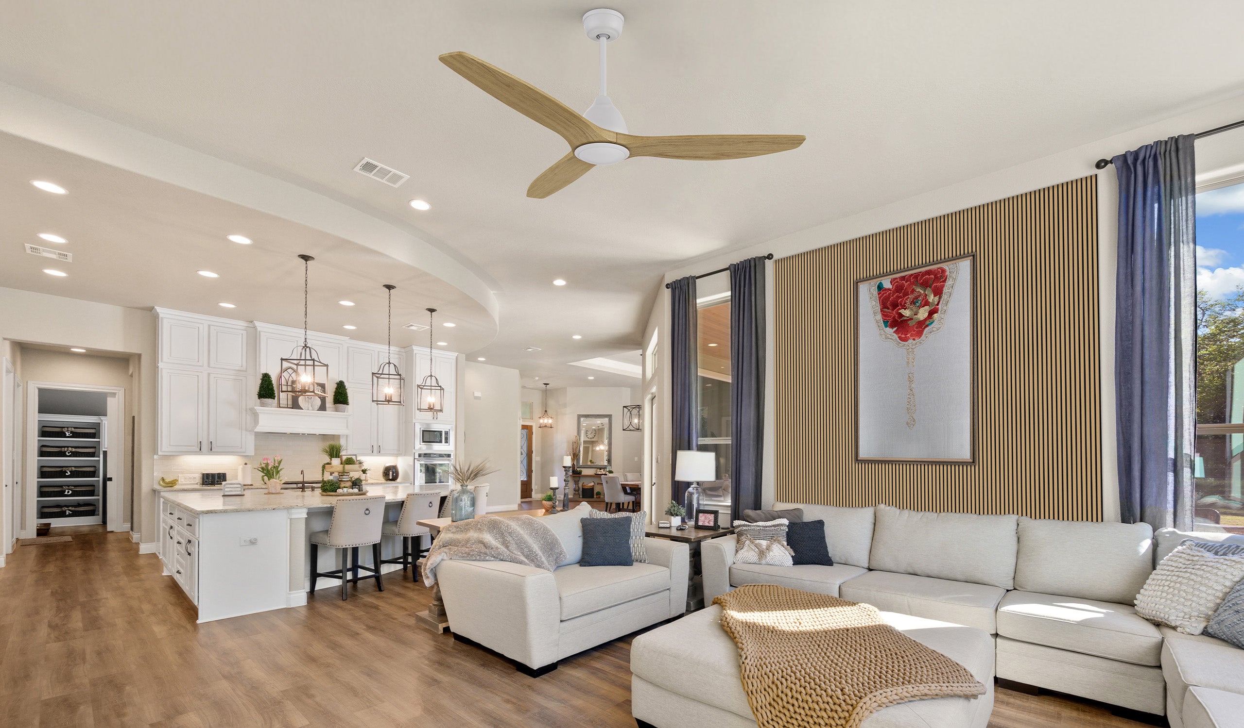 Spacious open-concept living room and kitchen featuring a modern wooden-blade ceiling fan with a white motor housing.  The space is designed with warm wood flooring, a white sectional sofa with textured pillows, and a cozy throw blanket.  A wooden accent wall with vertical slats complements the neutral decor, while pendant lights illuminate the kitchen island with bar seating.  Large windows with navy curtains allow natural light to brighten the space, creating a welcoming and stylish ambiance.