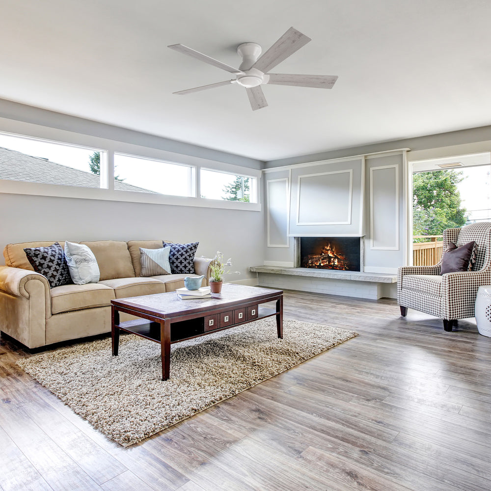 A modern living room featuring a Vetric 52 inch Low Profile Ceiling Fan with Remote No Light, positioned centrally on the white ceiling. The room includes a beige sofa with patterned pillows, a dark wood coffee table on a shag rug, a fireplace with a light gray mantel, and large windows offering a view of trees. 