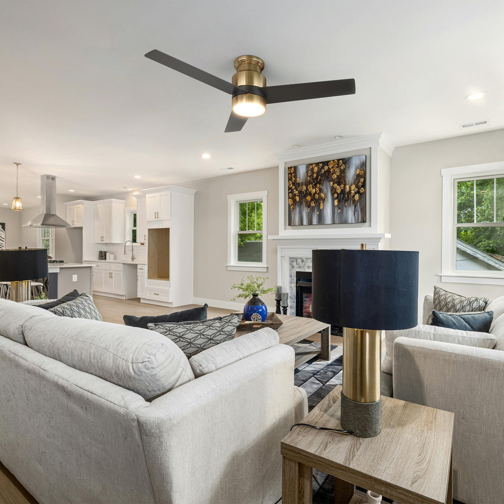 An open-concept living room and kitchen featuring a light grey sectional sofa with throw pillows, a wooden coffee table, and a matching side table with a gold-based lamp and dark blue shade. The room includes a fireplace with abstract artwork above it, and is illuminated by a flush mount ceiling fan in black and gold with an energy-efficient LED light kit, all styled in a modern farmhouse aesthetic with a neutral color palette. 