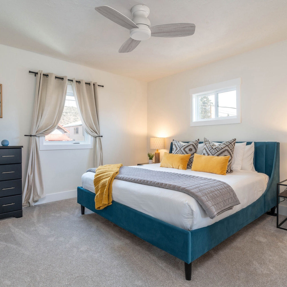A white, flush-mount ceiling fan without  light fixture is visible in a bedroom with a blue velvet bed frame, yellow and gray bedding, and a black dresser. The room also features two windows with light-colored curtains and a gray carpet.  