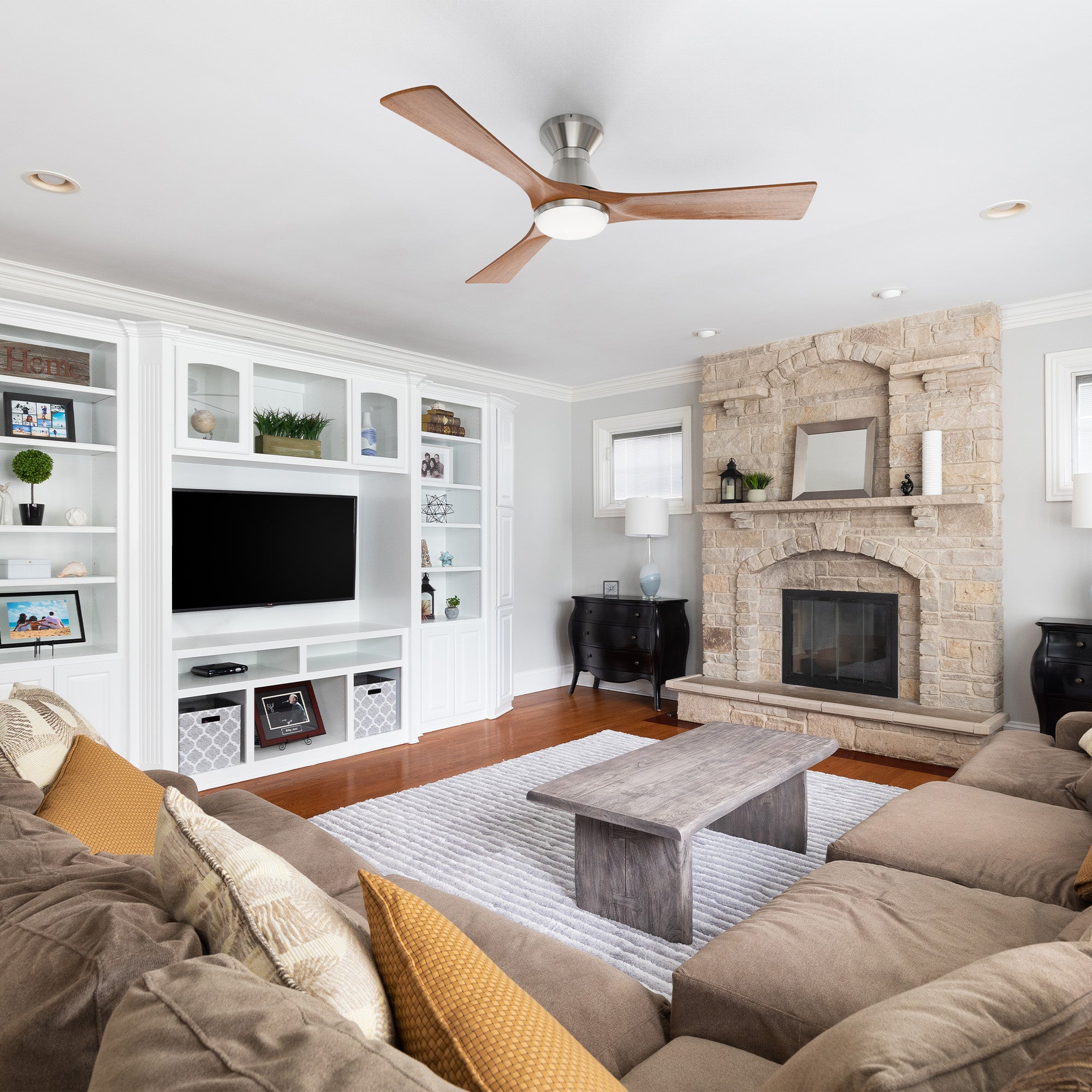 A modern living room with a brown sectional sofa, wooden coffee table, stone fireplace, built-in white shelves and TV, wood floor, gray rug, and a rustic farmhouse chic vibe. A low-profile ceiling fan with 3 blades completes the cozy Scandinavian-inspired space. 