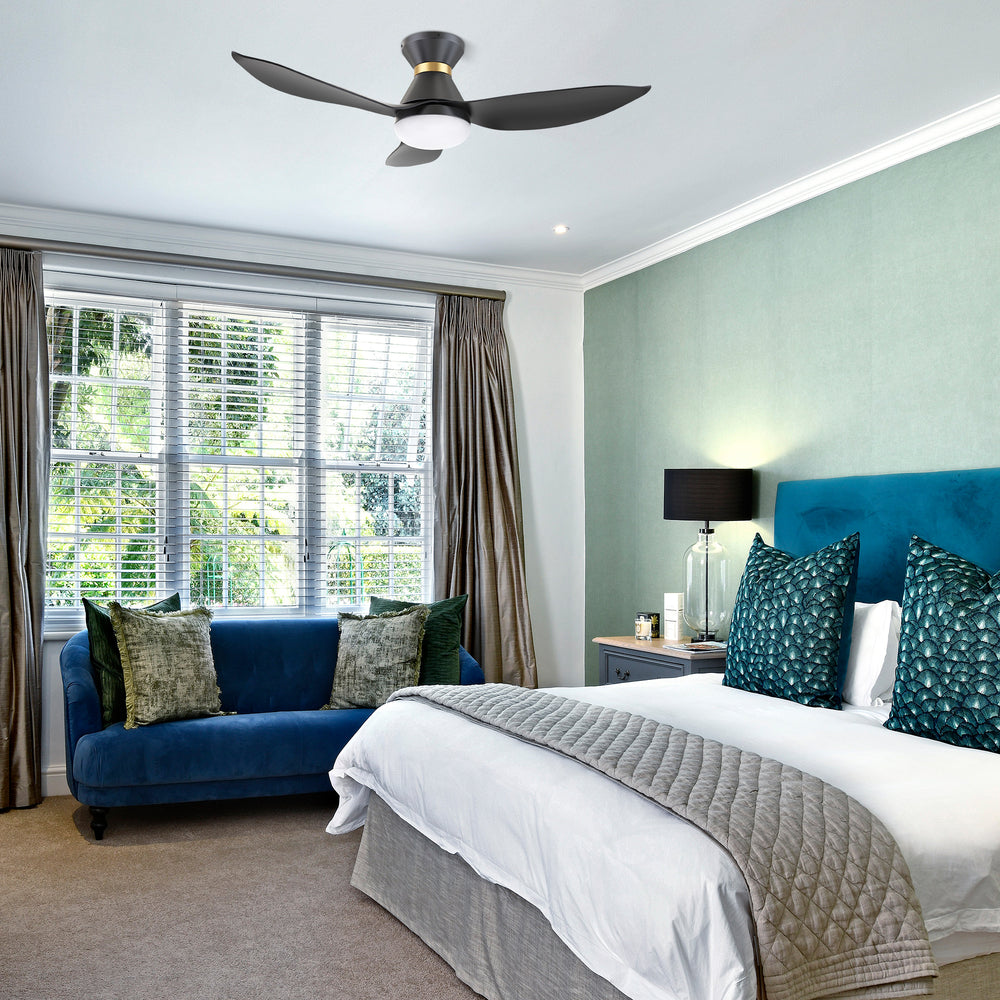 A modern bedroom featuring a 45-inch Joliet flush-mount smart ceiling fan in black and gold with three blades and an integrated light, mounted on a white ceiling. The room has a teal upholstered bed with white bedding and gray throw, teal and green cushions, a blue velvet sofa with green cushions, a nightstand with a lamp and decor, large windows with blinds and curtains, and a green wall with a beige carpeted floor. 