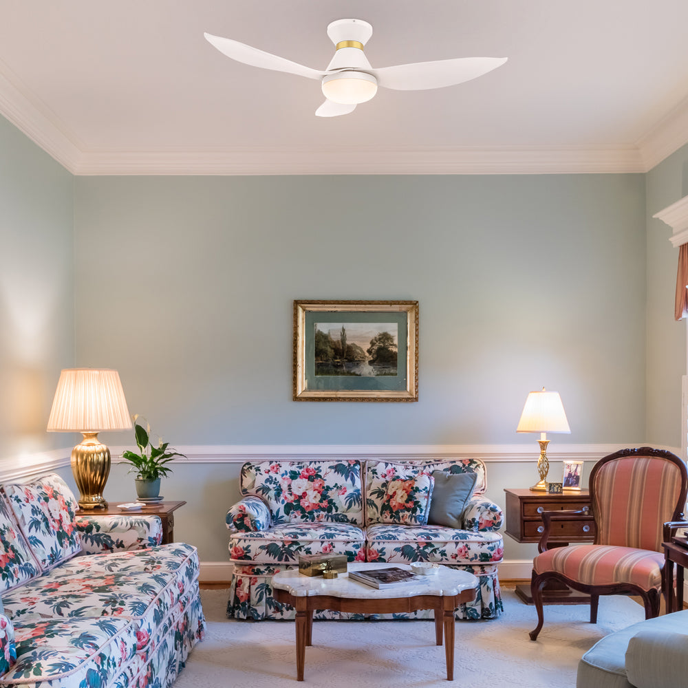 A cozy living room with a 45-inch Joliet flush-mount smart ceiling fan in white and gold with an integrated light, mounted on a white ceiling. The room features two floral-patterned sofas, a striped armchair, a wooden coffee table with a marble top, side tables with gold lamps, a framed picture on the wall, and a plant, all set against light green walls and a carpeted floor. 